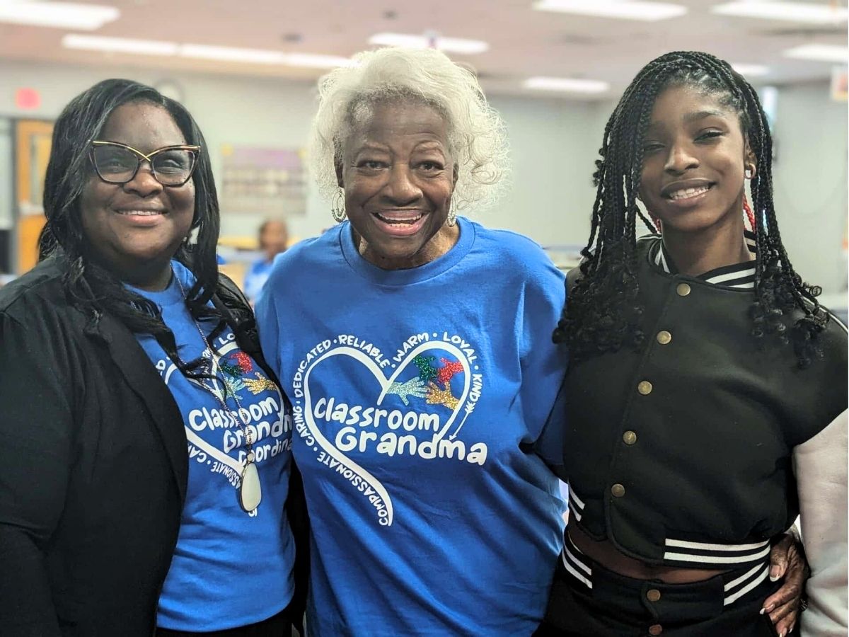 Assistant Principal Sa’Tonya Brascom, Classroom Grandparent Grandma Woody, and student Harmoni smiling together at school.